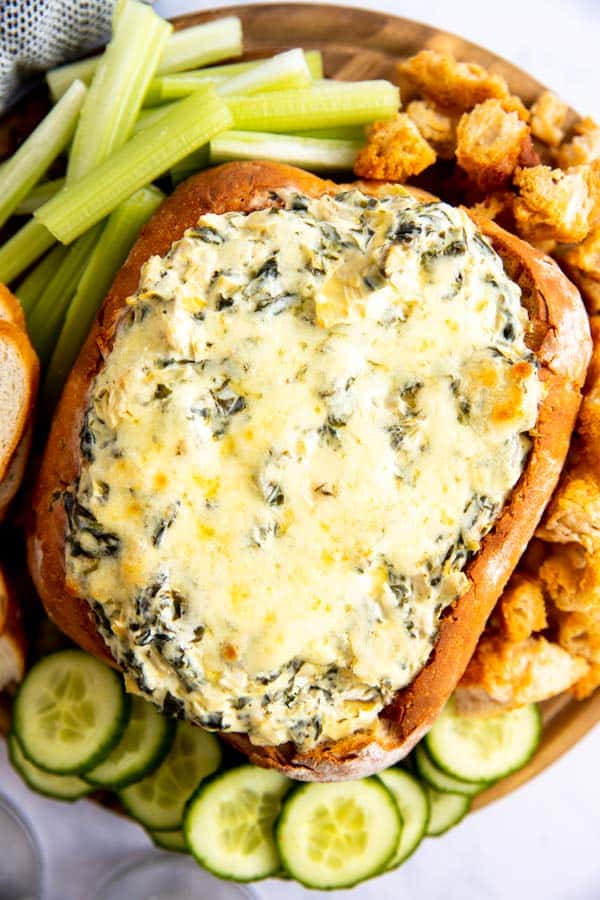 vertical image of wooden board with bread bowl filled with spinach artichoke dip, surrounded by vegetables and bread