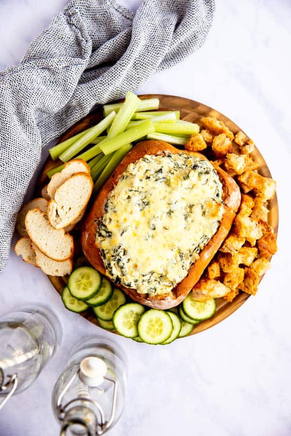 white marble surface with a wooden appetizer tray with spinach artichoke bread bowl dip