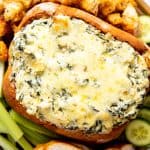 wooden board with bread bowl filled with spinach artichoke dip, surrounded by vegetables and bread