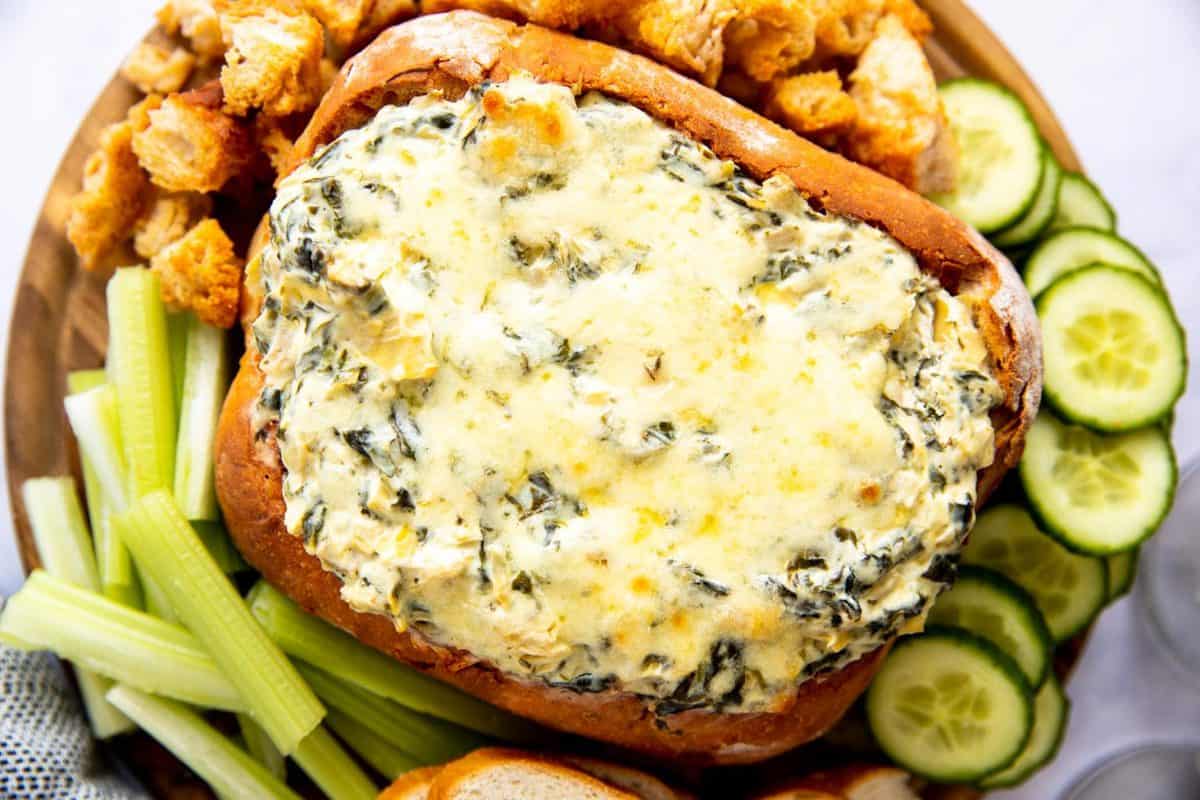 wooden board with bread bowl filled with spinach artichoke dip, surrounded by vegetables and bread
