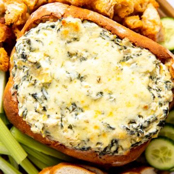 wooden board with bread bowl filled with spinach artichoke dip, surrounded by vegetables and bread