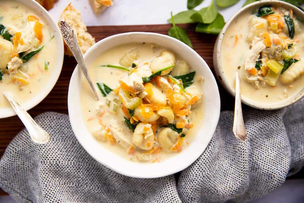 three white bowls with chicken gnocchi soup on a wooden board