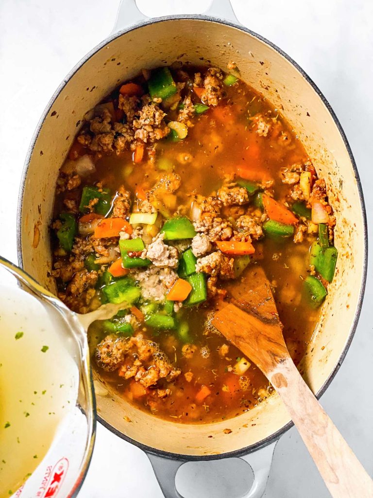 broth pouring from glass measuring cup over sausage and vegetables in white Dutch oven