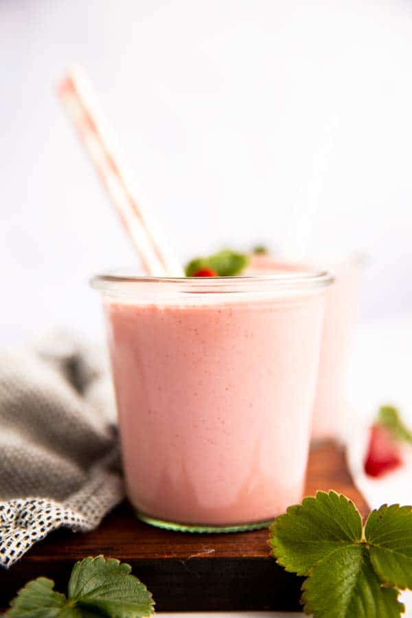 two glasses of strawberry smoothie with straws on a wooden board with strawberry leaves