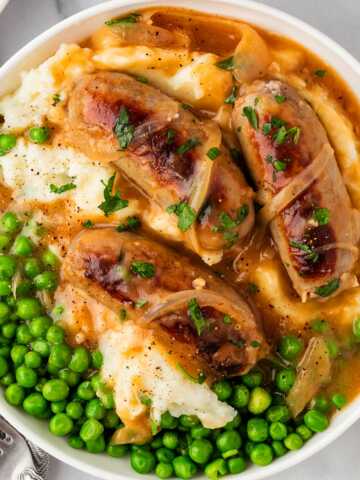 overhead view of mashed potatoes, peas and sausages with onion gravy in white bowl