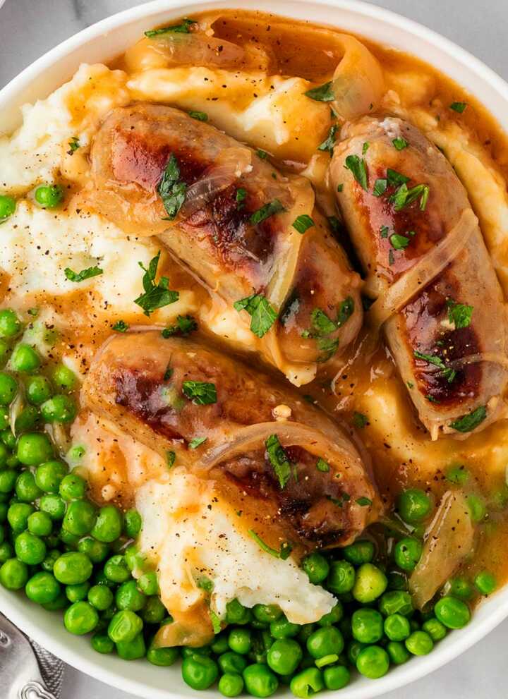 overhead view of mashed potatoes, peas and sausages with onion gravy in white bowl