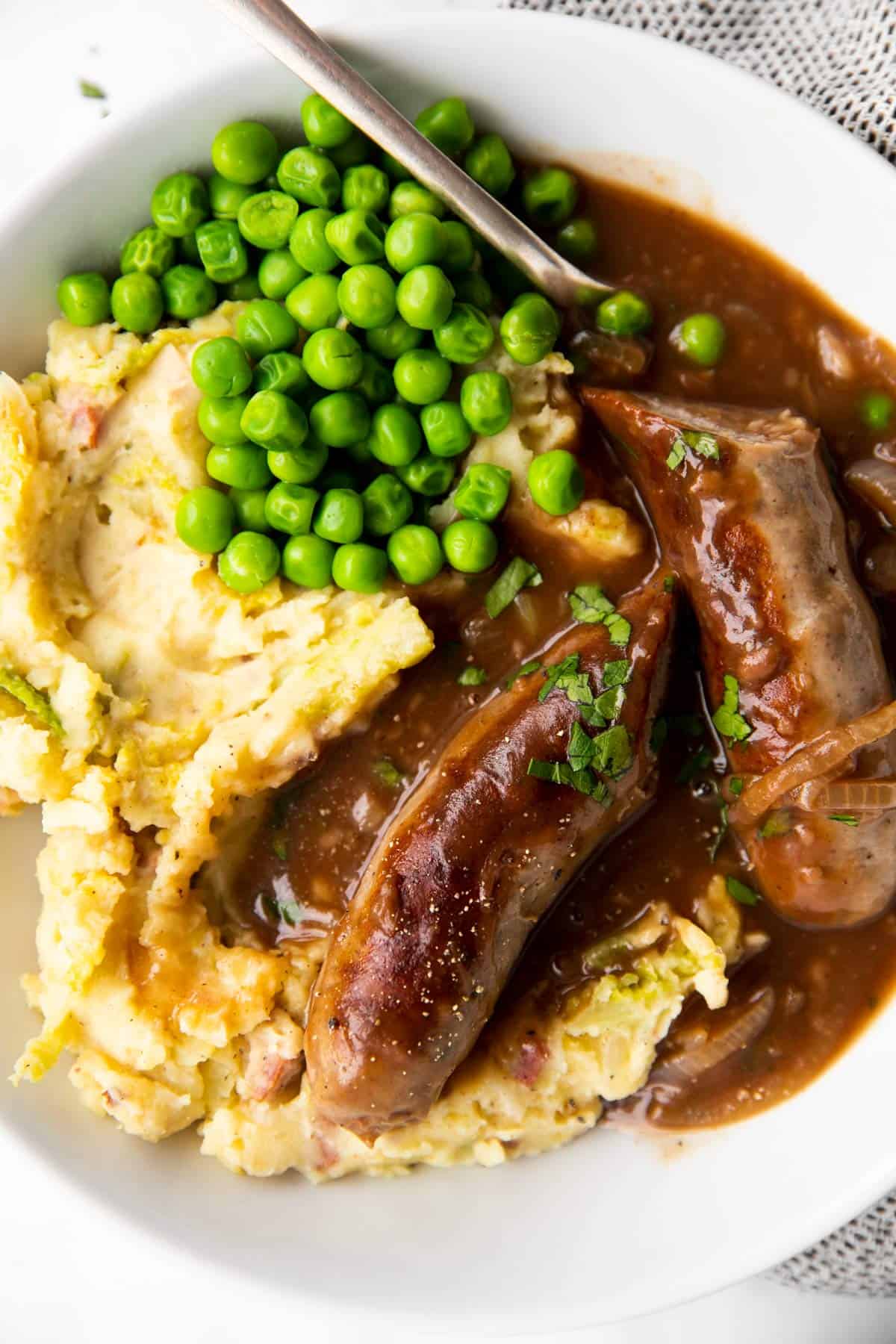 close up photo of sausages on a plate with mashed potatoes, peas and onion gravy