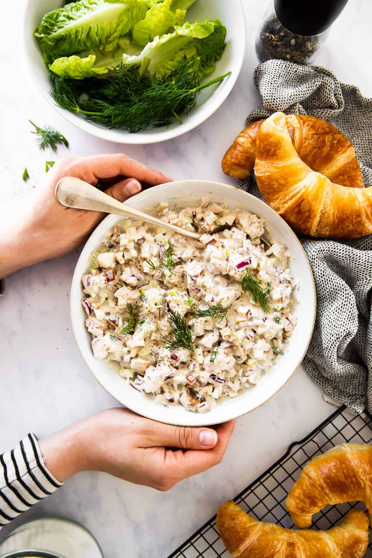 top down view on female hands holding white bowl with chicken salad next to croissants