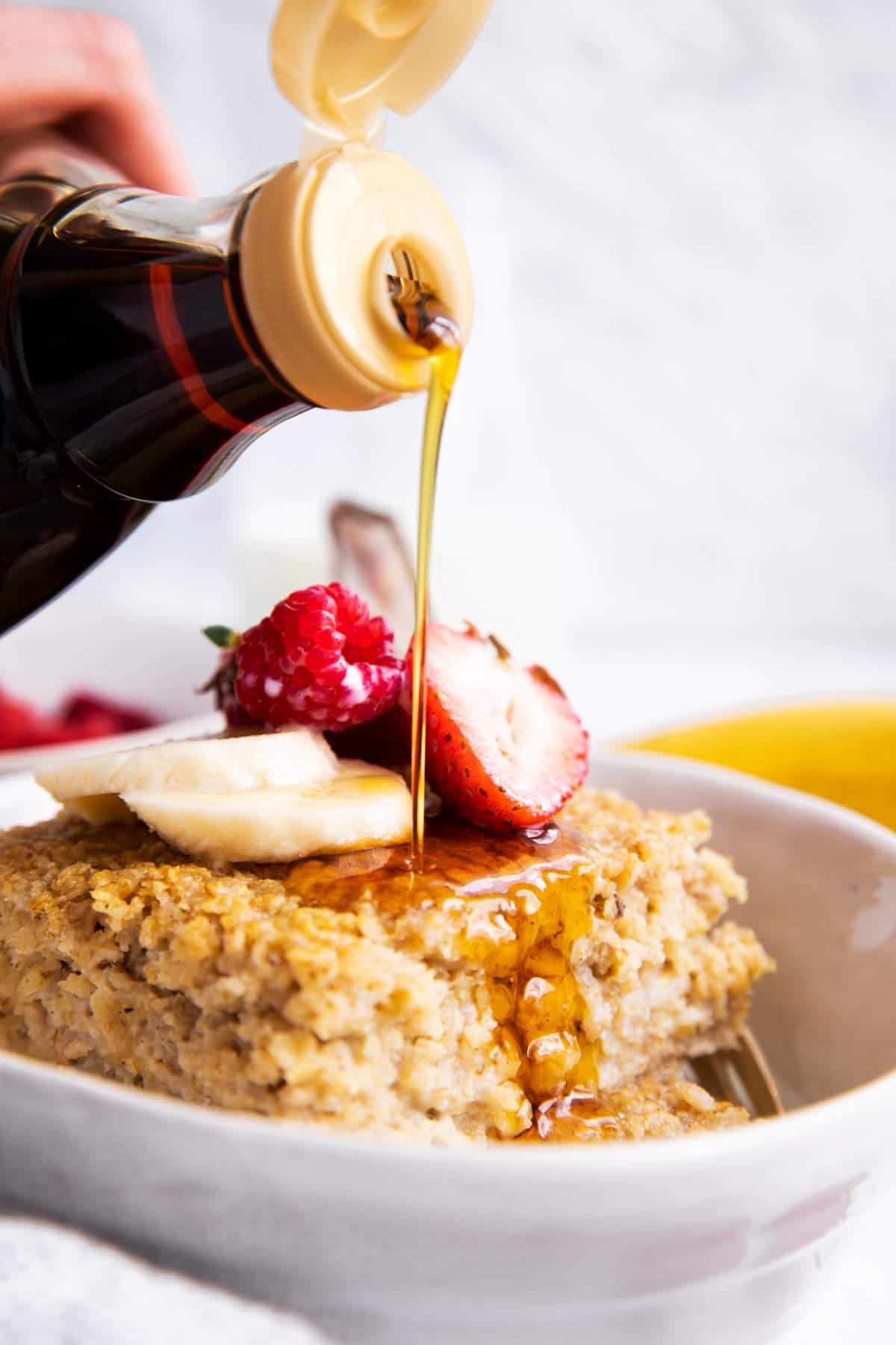 pouring maple syrup over baked oatmeal in a white bowl with fresh fruit