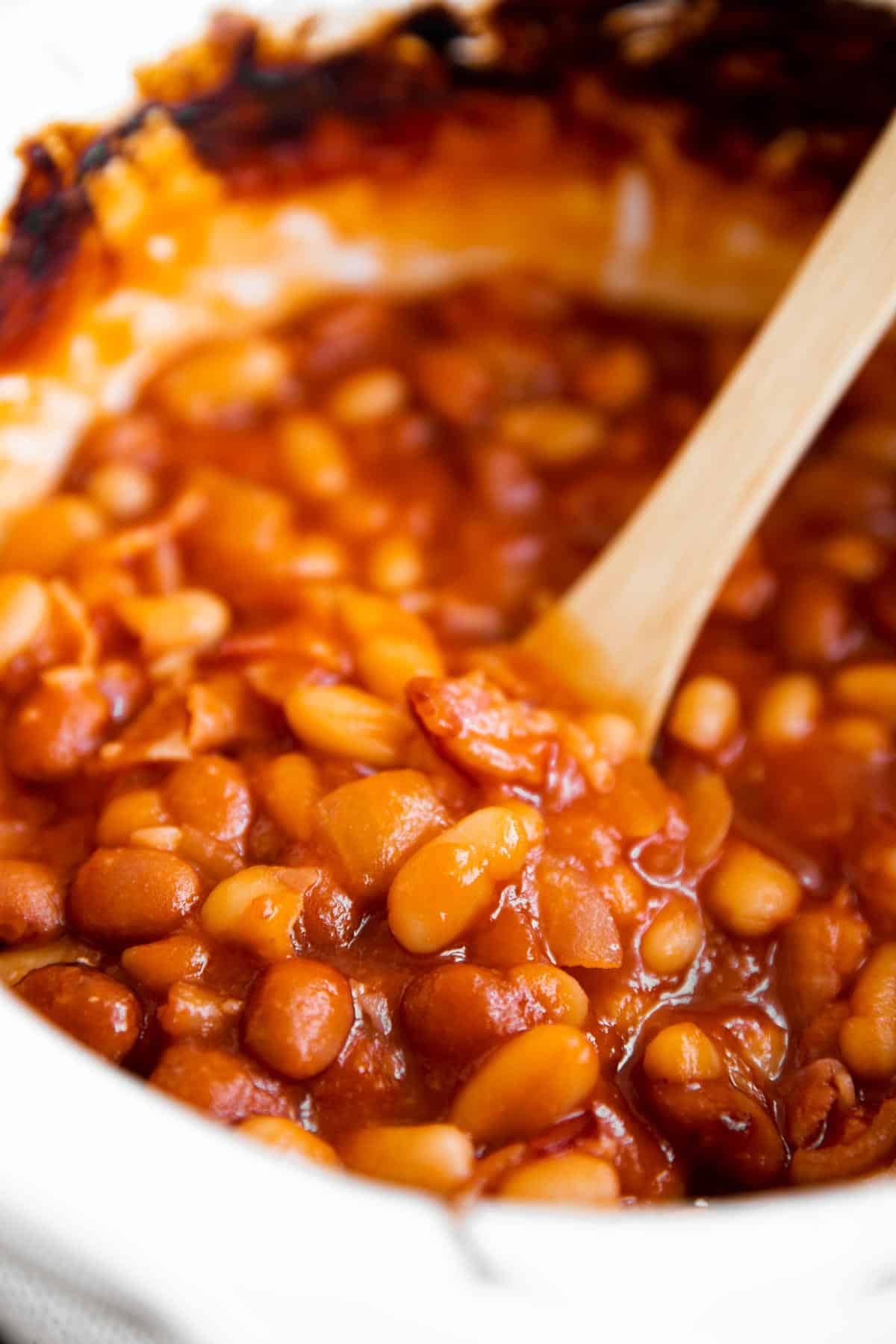 close up photo of homemade baked beans in a white crock with a wooden spoon