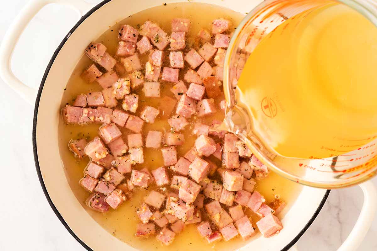 Pouring broth into skillet with ham cubes.