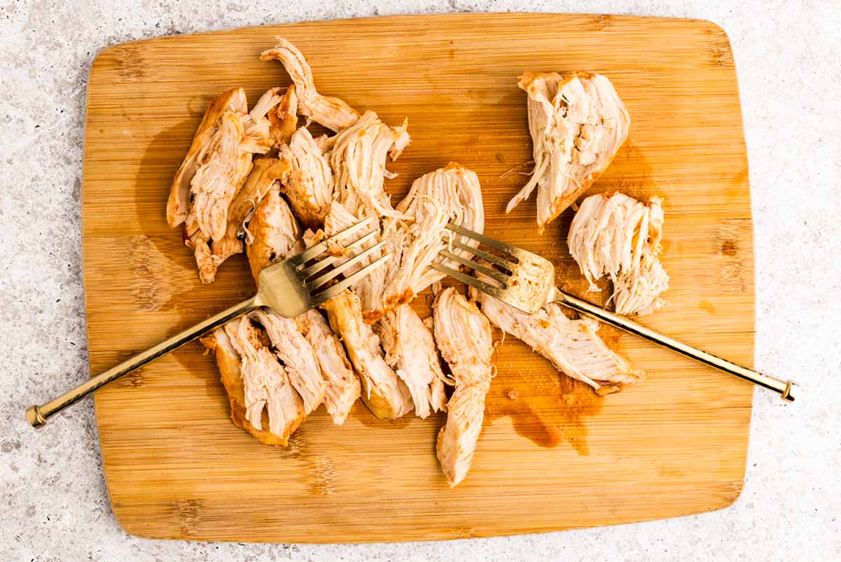 Pulling cooked chicken breast into chunks on a wooden cutting board using forks.