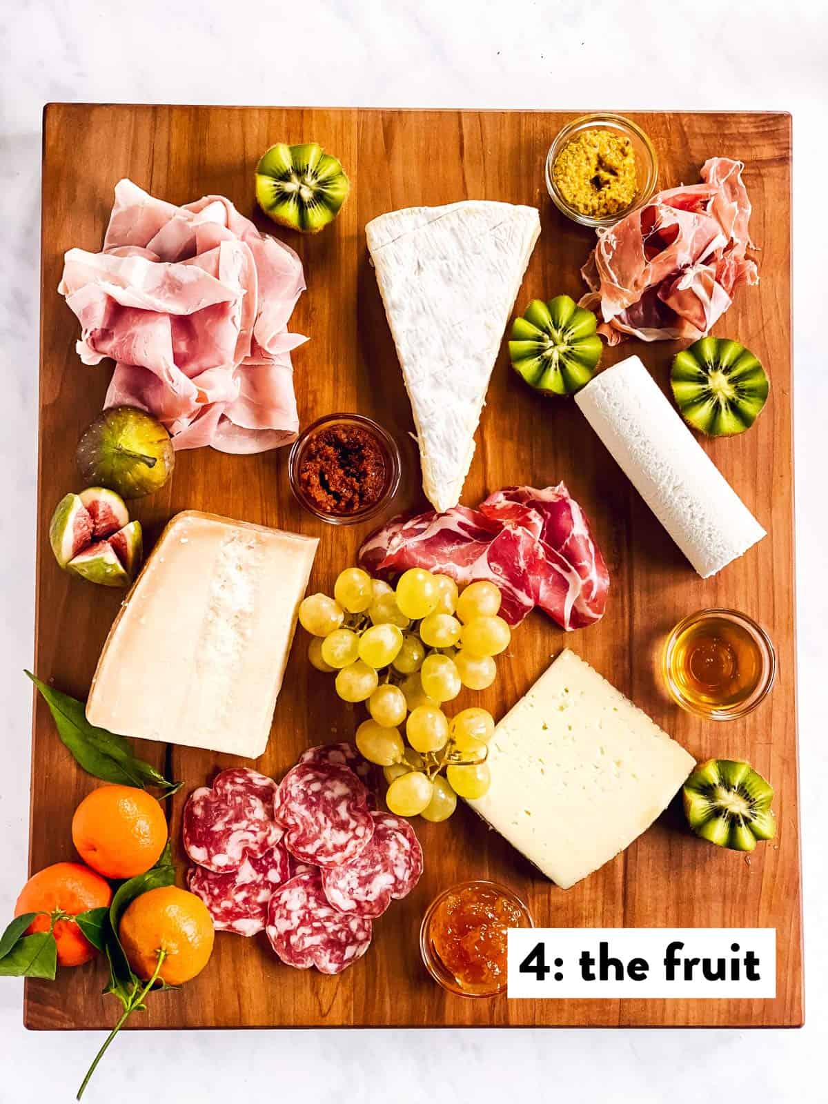 four types of cheese, cold cuts, spreads and fruit on a wooden board