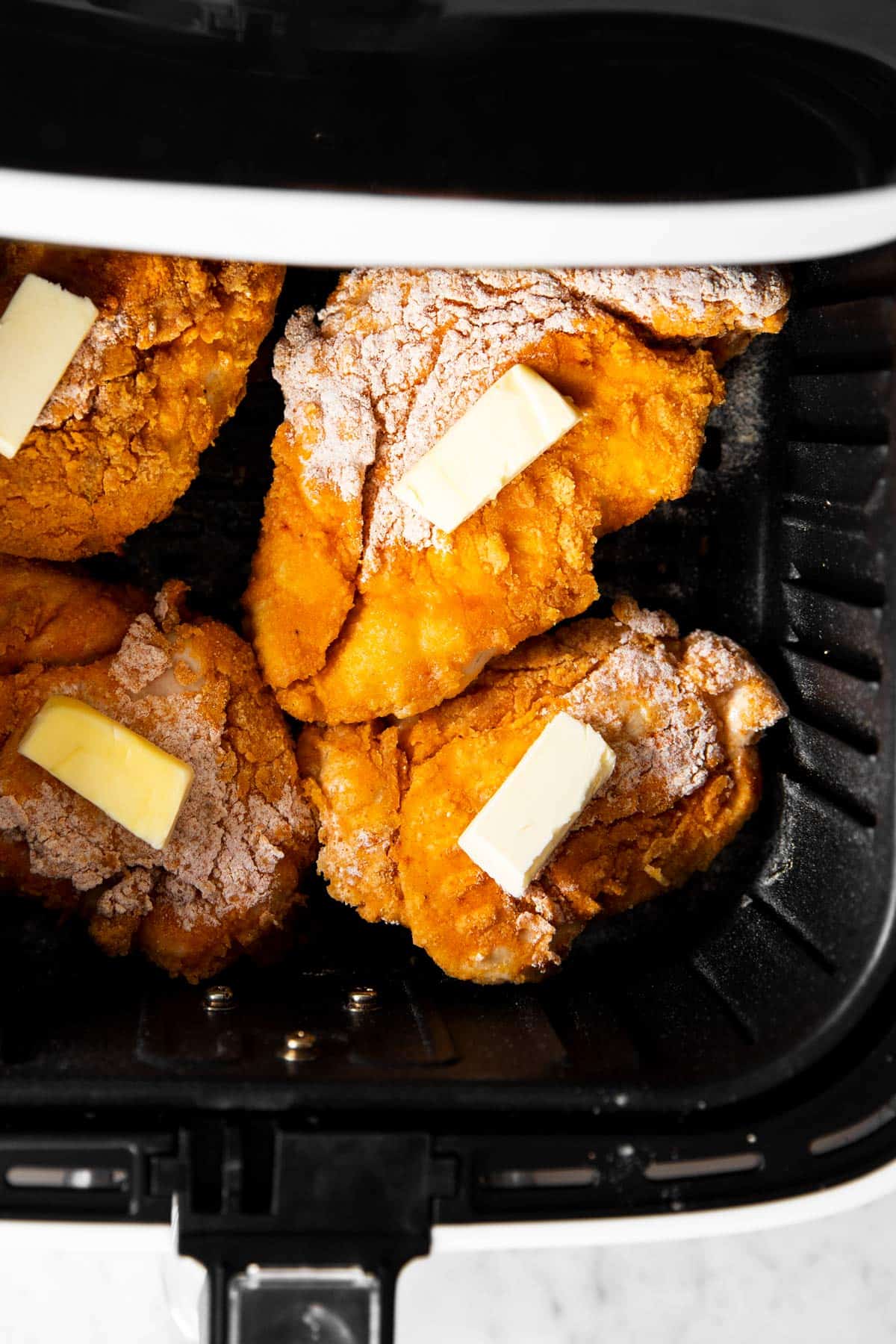 half cooked fried chicken in air fryer basket topped with butter