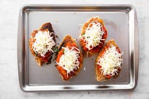 four breaded chicken breasts topped with marinara sauce, herbs and cheese on sheet pan