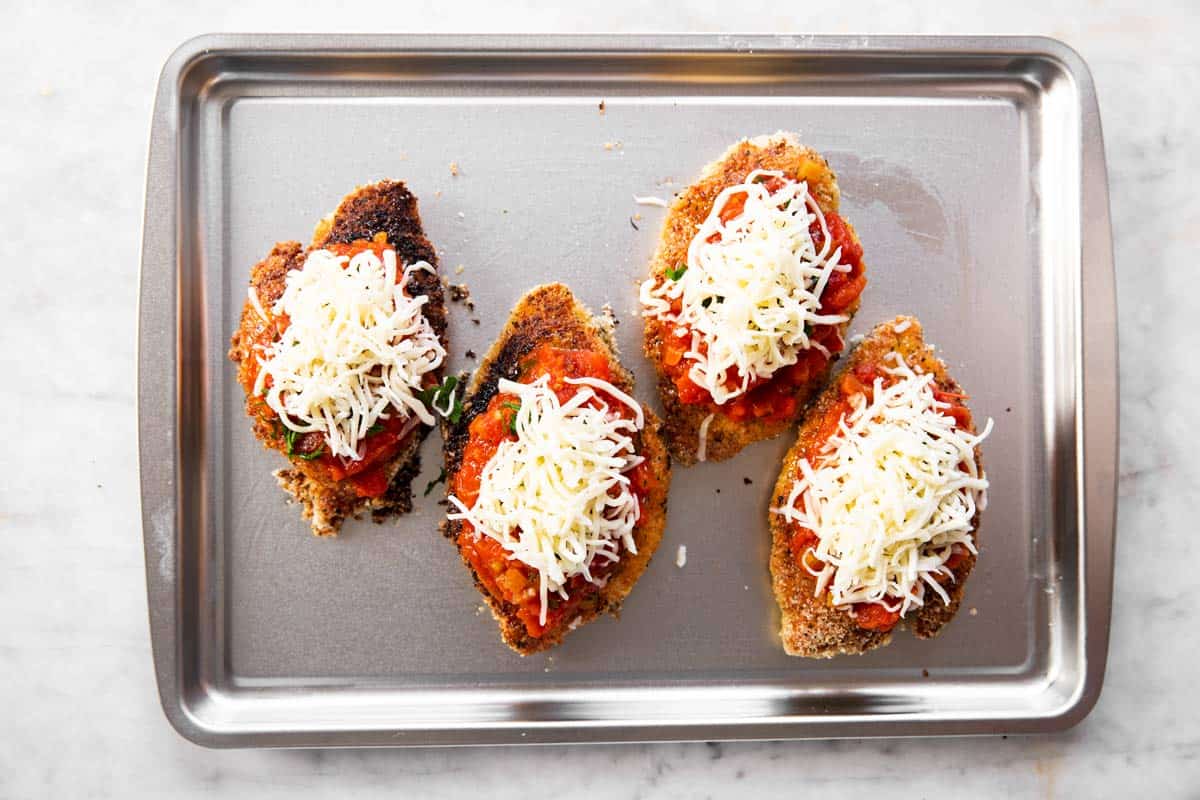 four breaded chicken breasts topped with marinara sauce, herbs and cheese on sheet pan