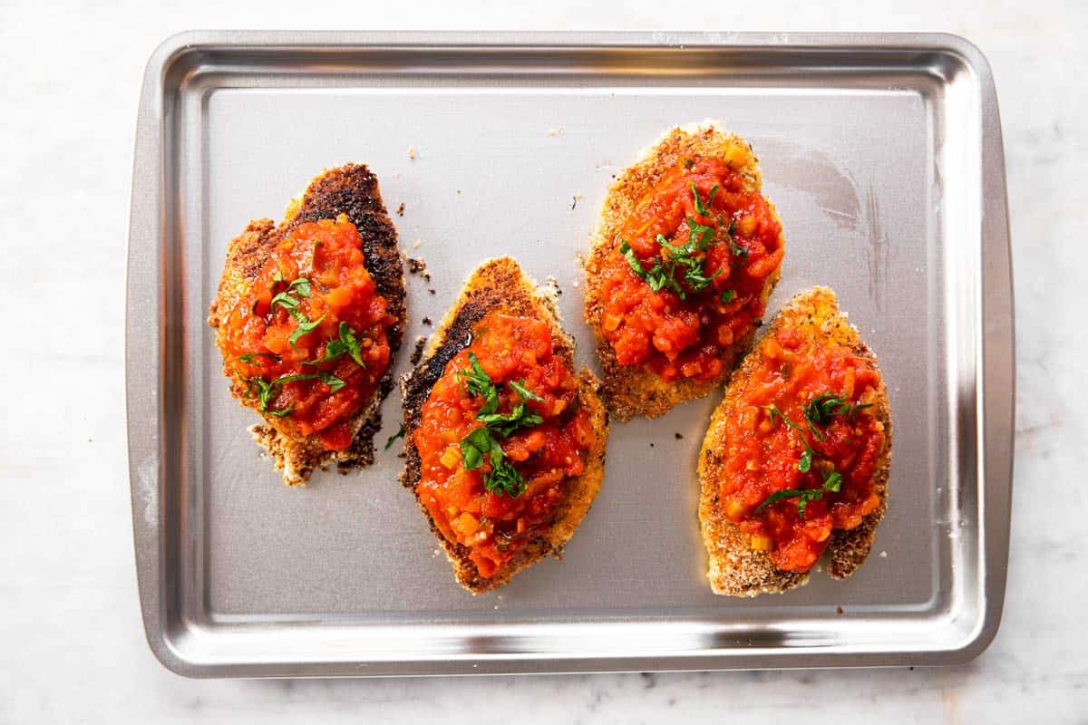 four breaded chicken breasts topped with marinara sauce and herbs on sheet pan