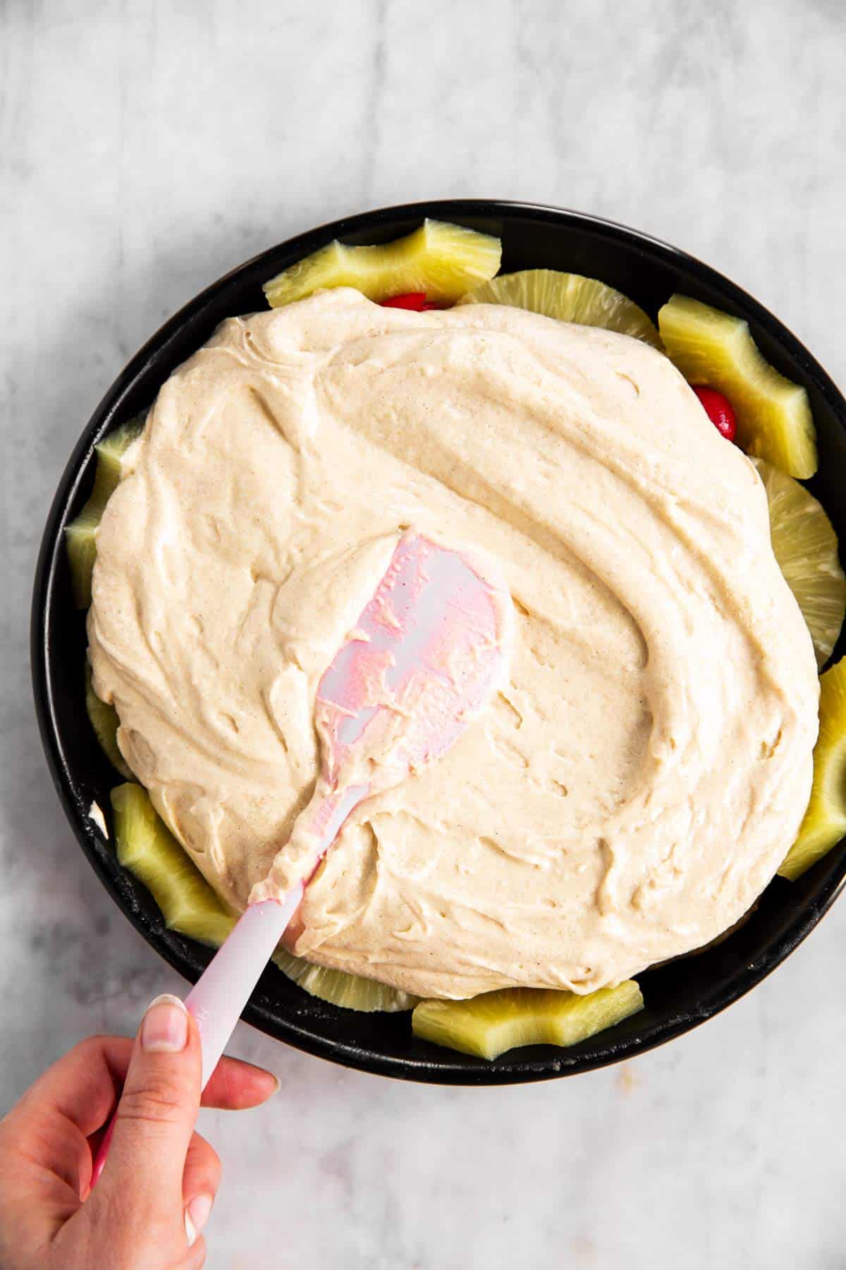 female hand spreading cake batter over pineapples and cherries in black pan