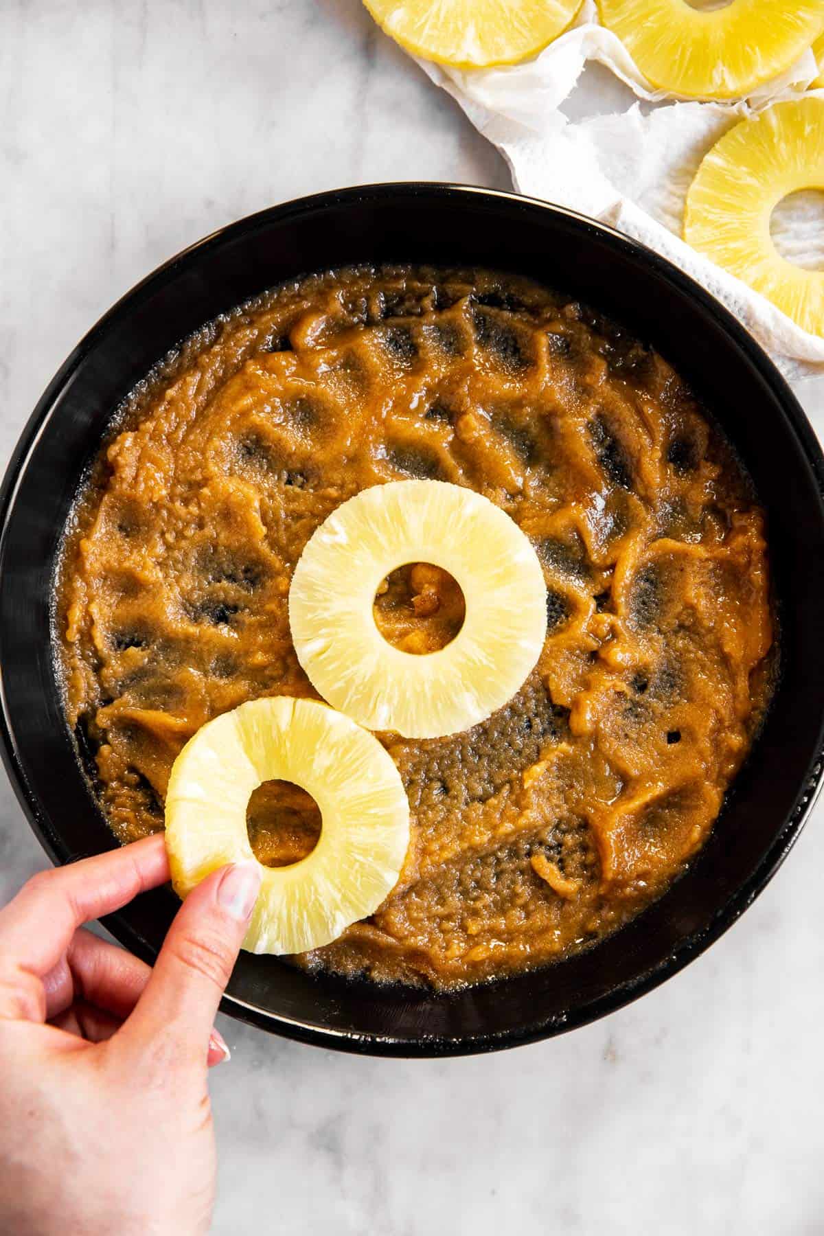female hand placing pineapple rings in black cake pan filled with brown sugar and butter paste