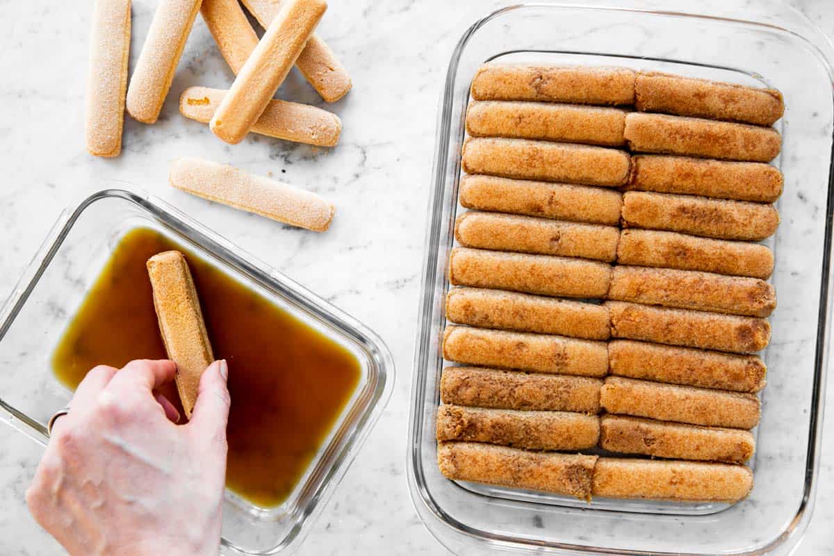 female hand dipping ladyfinger biscuit into coffee next to glass pan filled with dipped Savoiardi biscuits