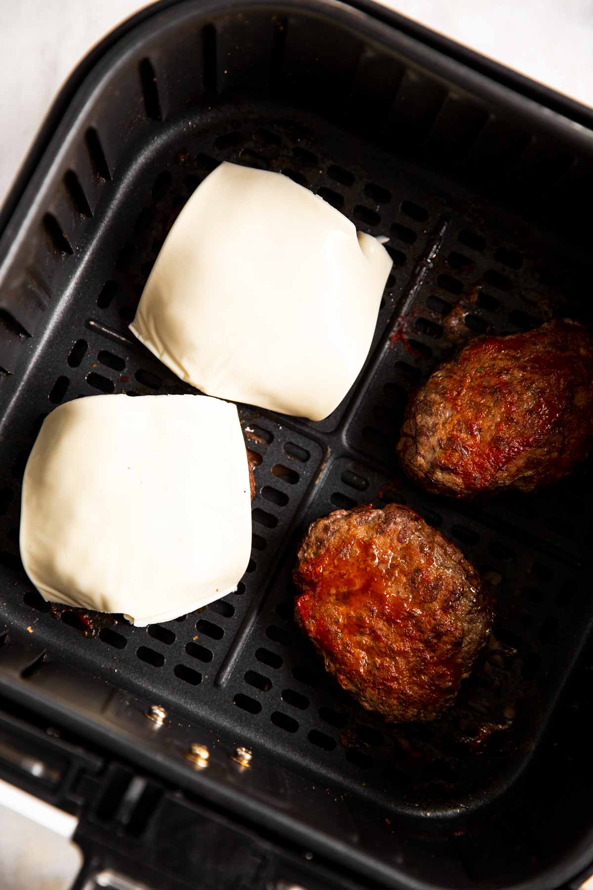 four burger patties in air fryer basket, two covered with slice of cheese