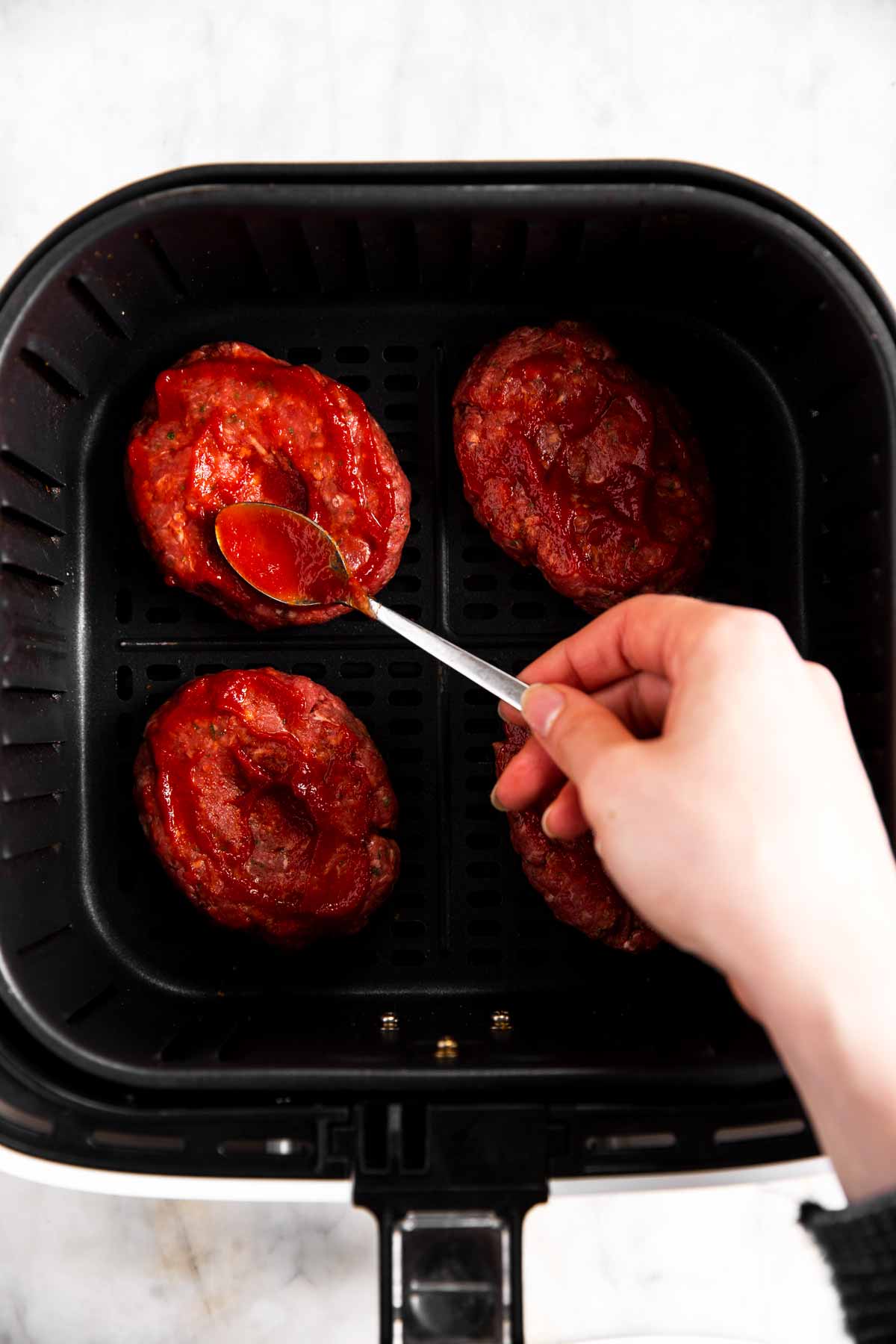 female hand brushing ketchup over uncooked burger patties in air fryer basket