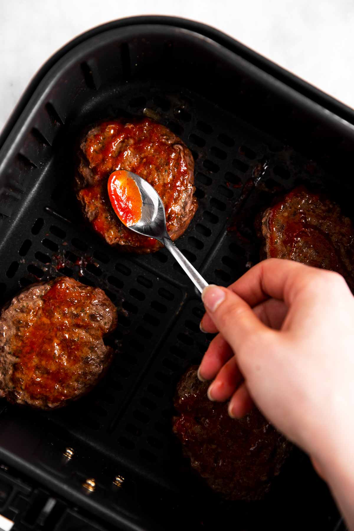 female hand spreading ketchup over cooked burger patty in air fryer basket