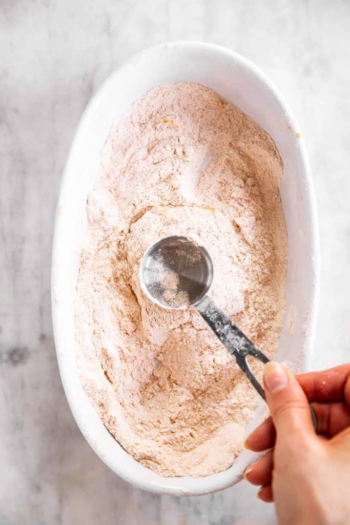 female hand pounding piece of breaded fish fillet with back of small measuring cup