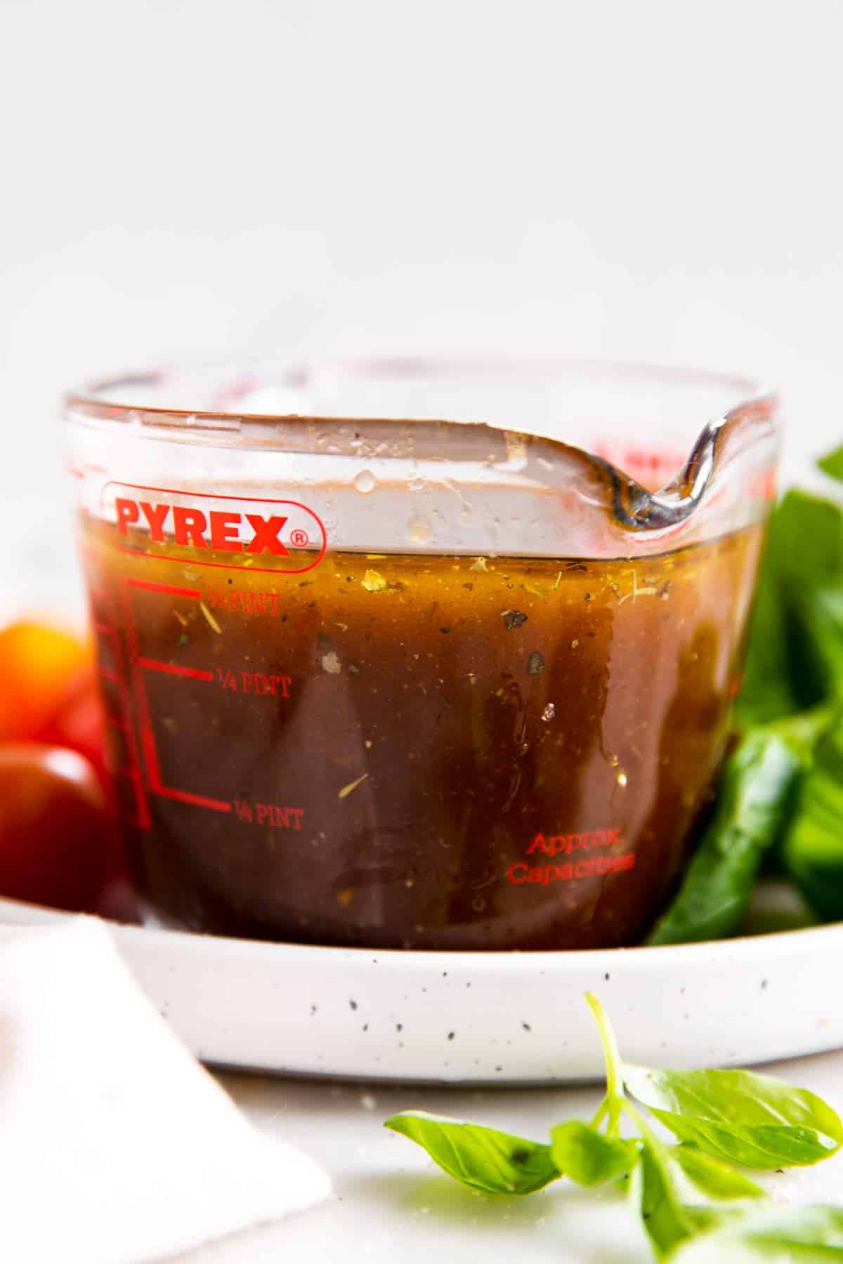 measuring jug with balsamic vinaigrette sitting on plate with basil and cherry tomatoes