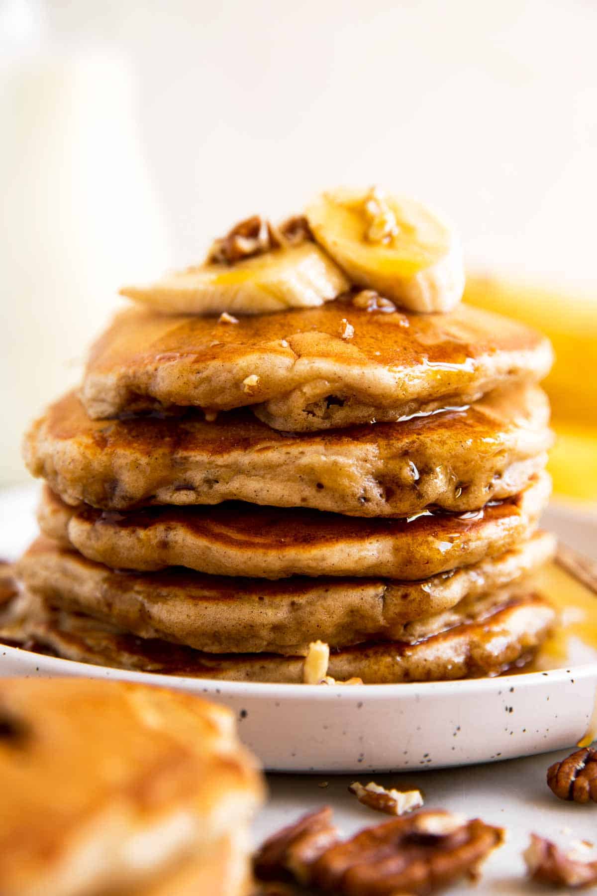 stack of banana pancakes on white plate with maple syrup, banana slices and pecans