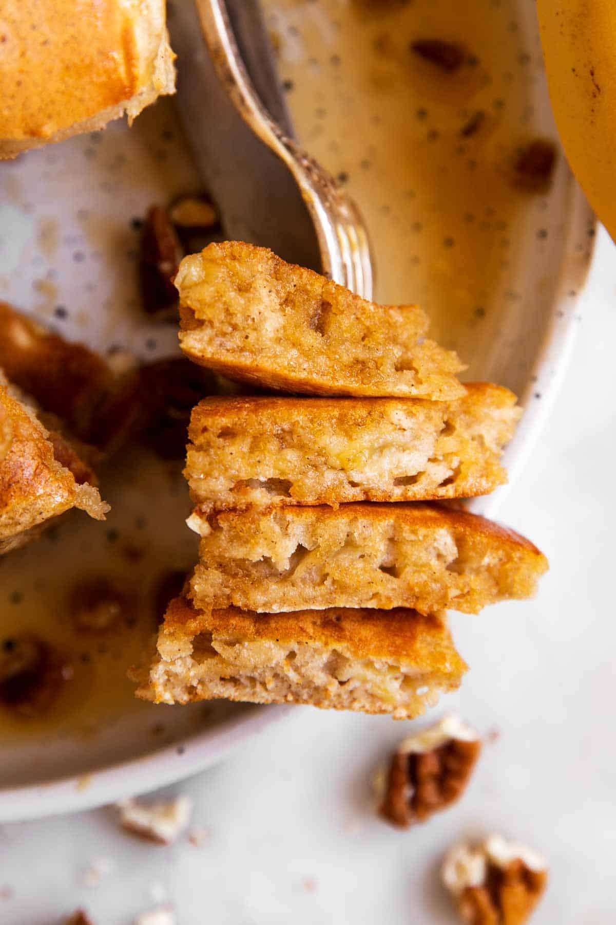 four pieces of banana pancake speared on fork, sitting on white plate with pecans and maple syrup