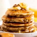 stack of banana pancakes on white plate with maple syrup, banana slices and pecans