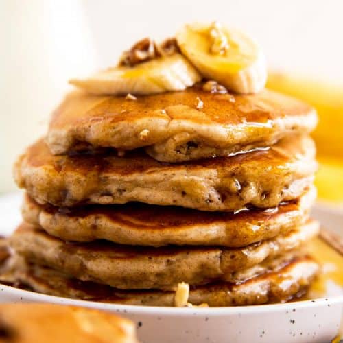 stack of banana pancakes on white plate with maple syrup, banana slices and pecans