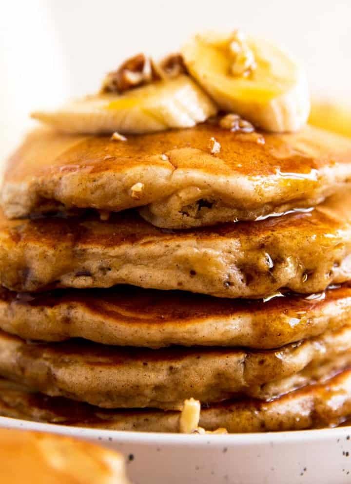 stack of banana pancakes on white plate with maple syrup, banana slices and pecans
