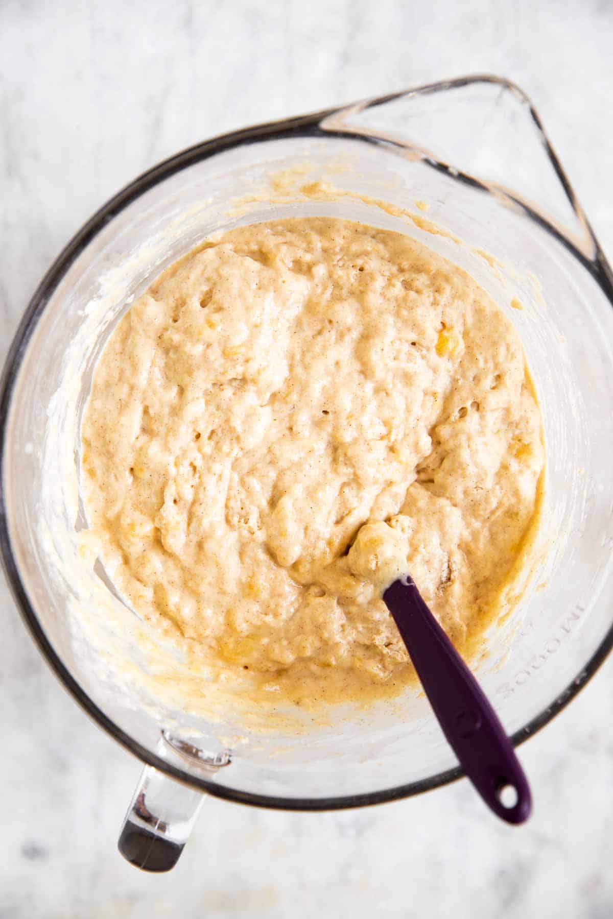 glass mixing bowl with banana pancake batter, with purple rubber spatula stuck in batter