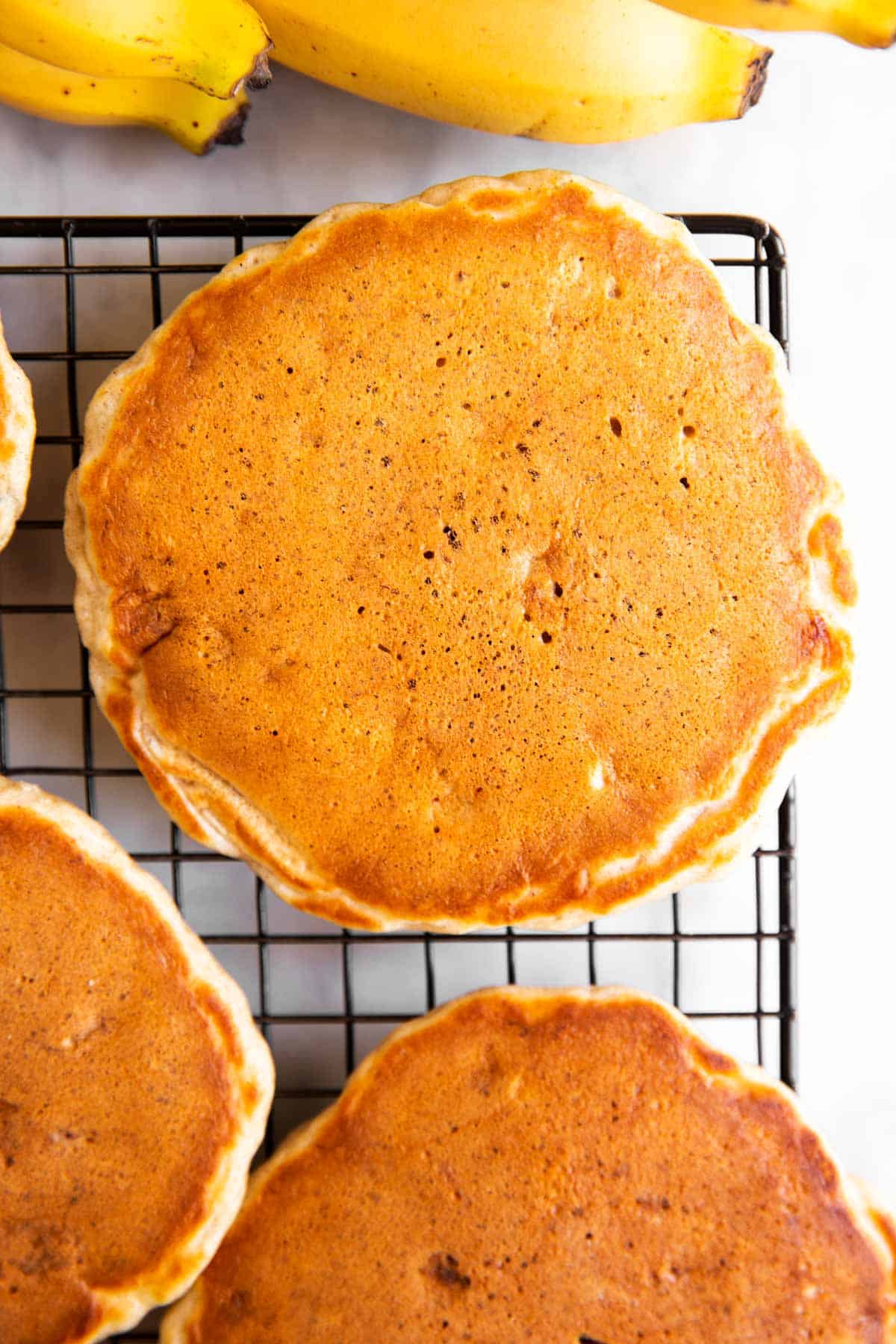 three cooked banana pancakes on cooling rack, next to three unpeeled bananas