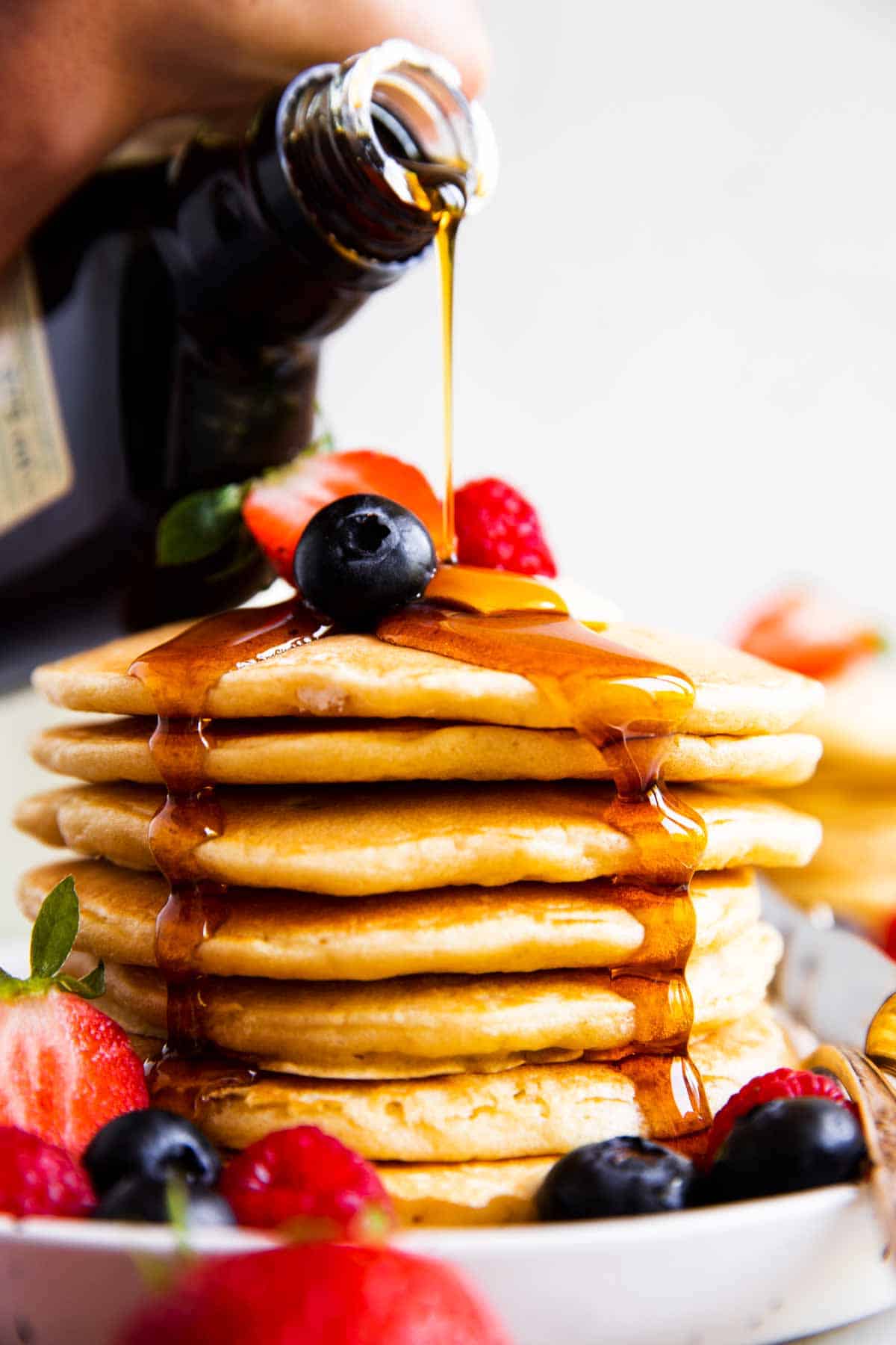 maple syrup pouring over stack of buttermilk pancakes with berries