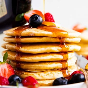 maple syrup pouring over stack of buttermilk pancakes with berries