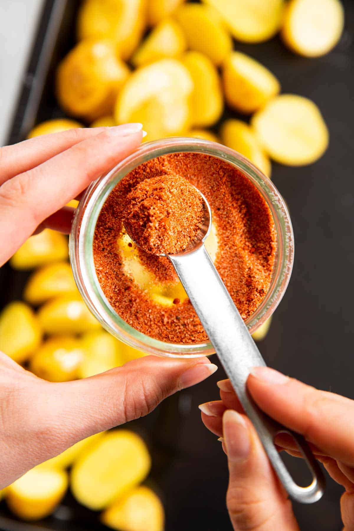 female hands scooping seasoning mix from a small glass jar with teaspoon measure