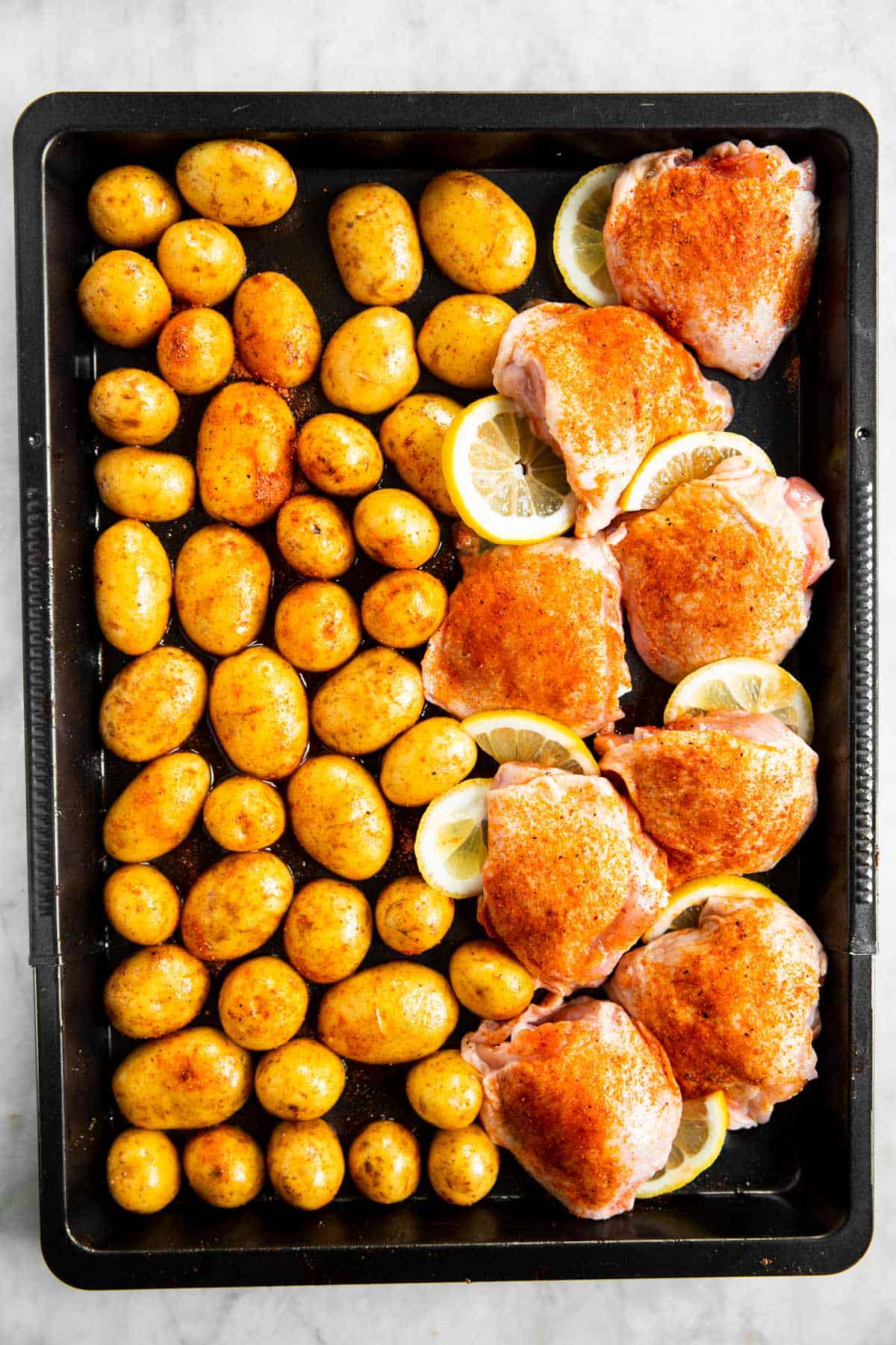 overhead view of seasoned potatoes and chicken thighs with lemon slices on black sheet pan