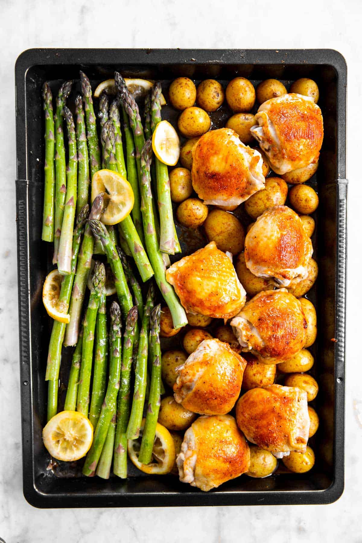 overhead view of half-roasted potatoes and chicken thighs with raw asparagus on black sheet pan