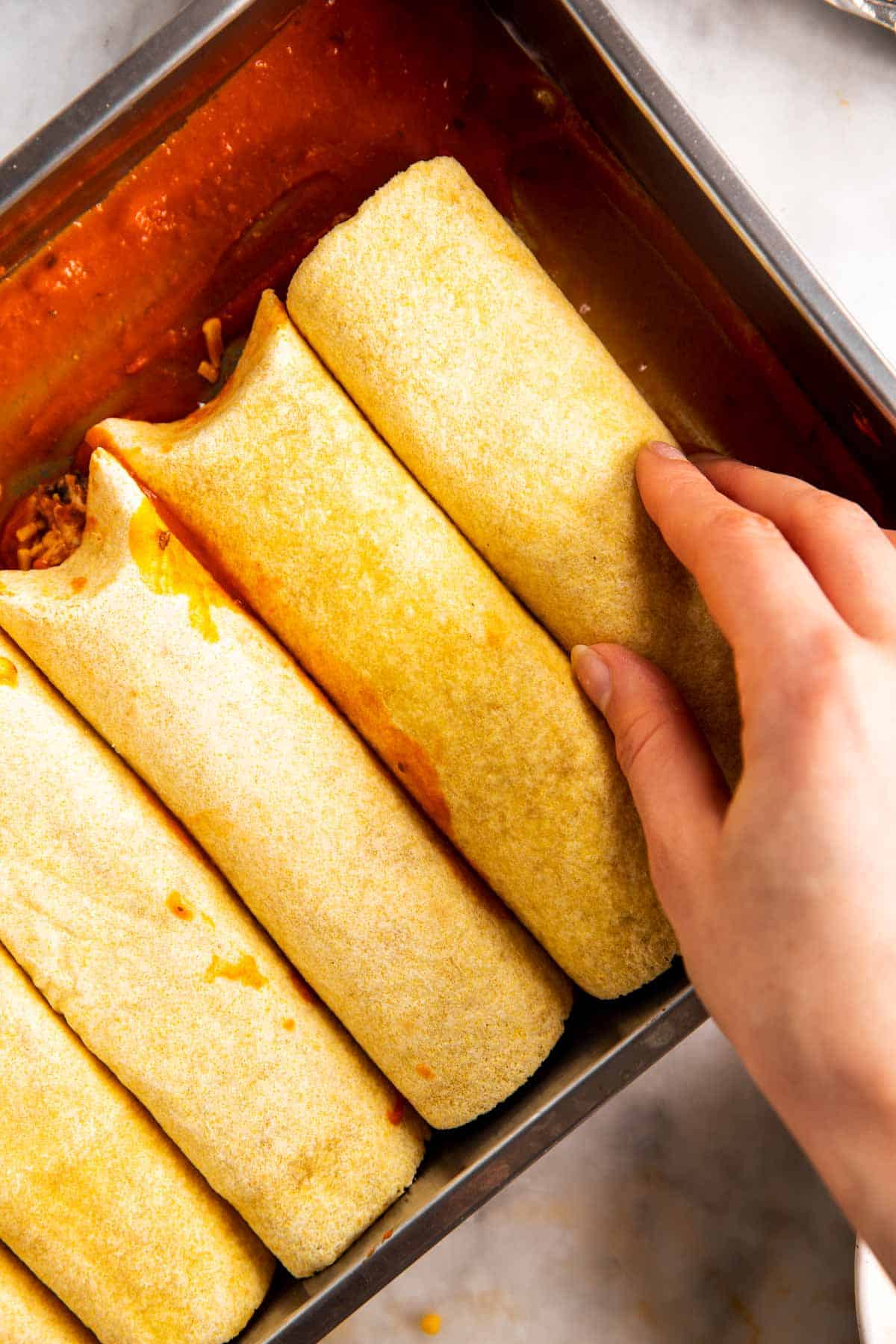 female hand placing folded corn tortilla inside casserole dish with other assembled chicken enchiladas