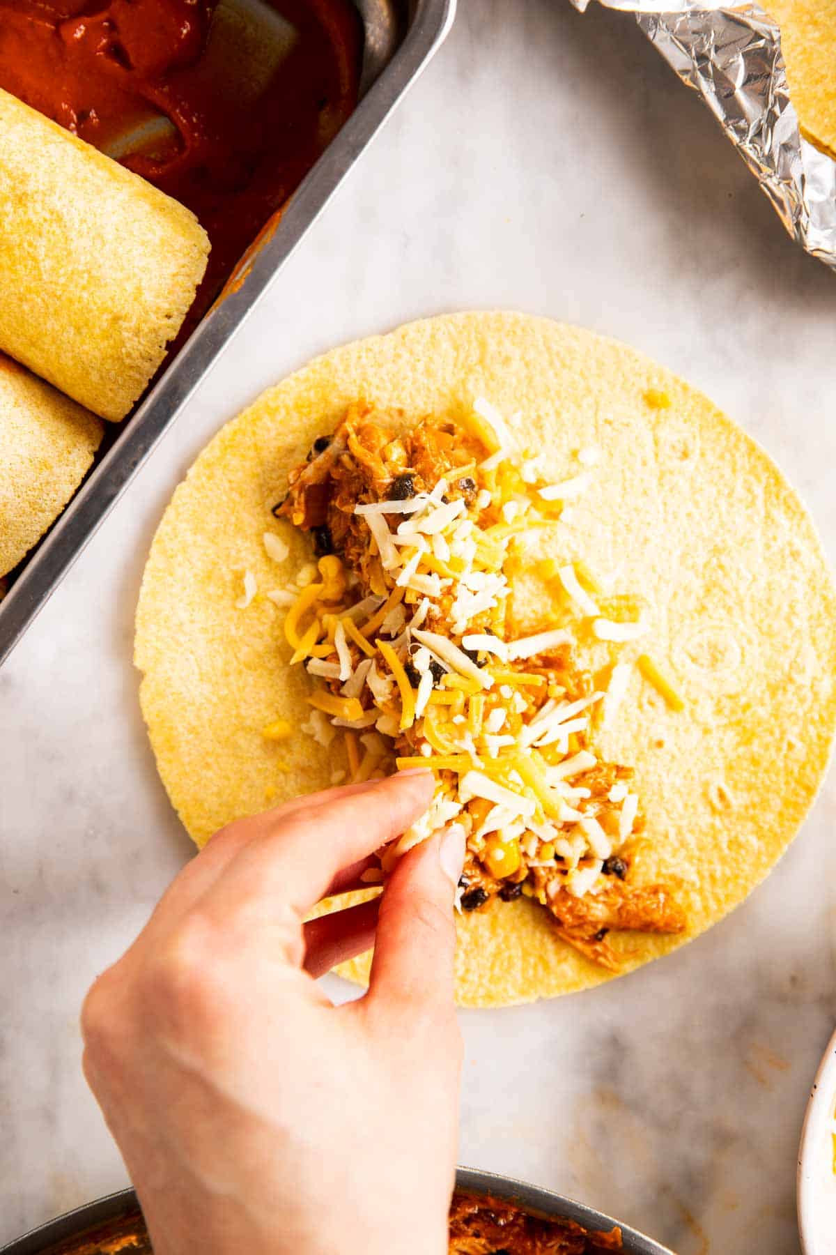 female hand sprinkling shredded cheese over chicken enchilada filling on corn tortilla