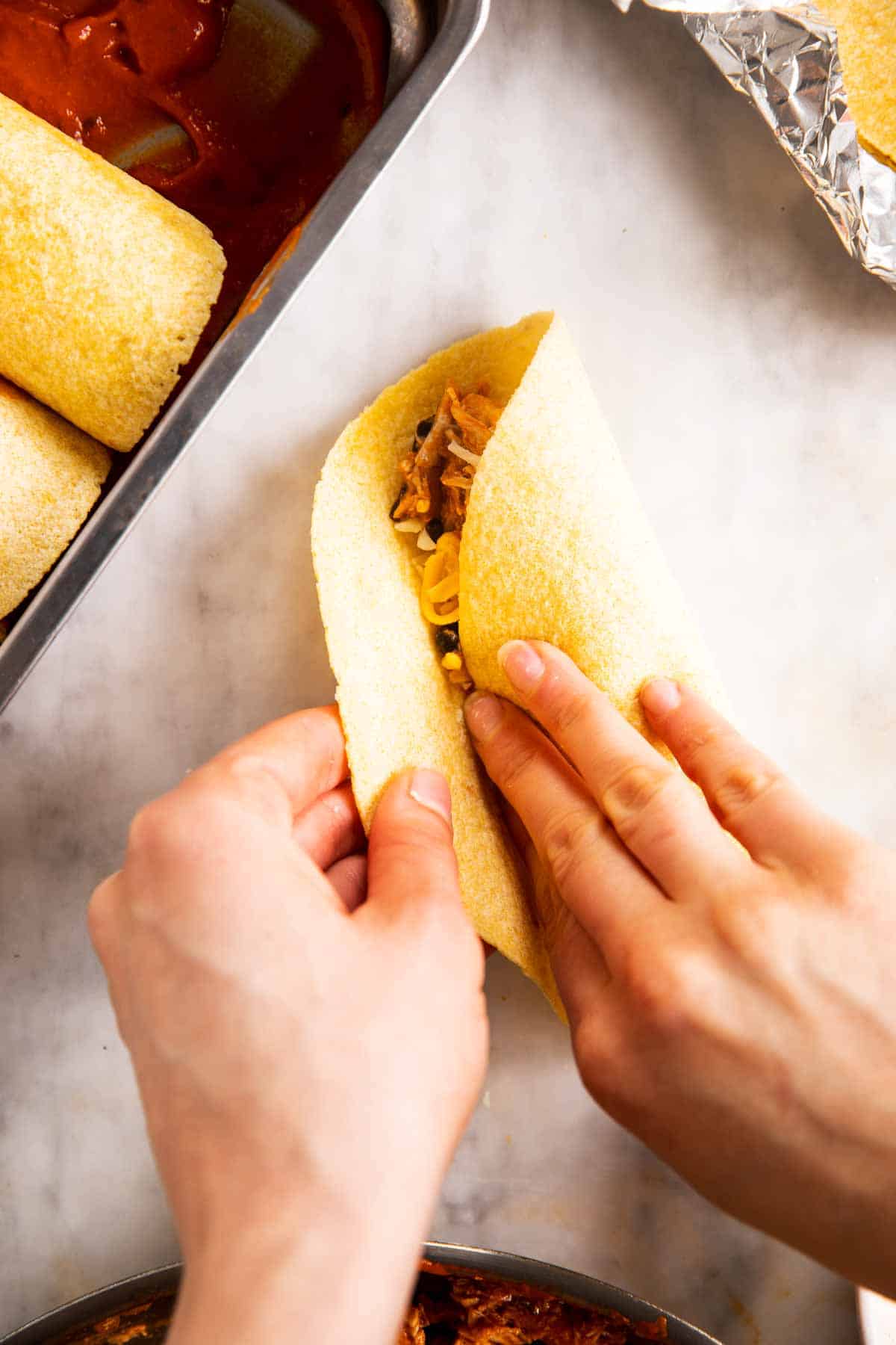 female hands folding corn tortilla over chicken enchilada filling