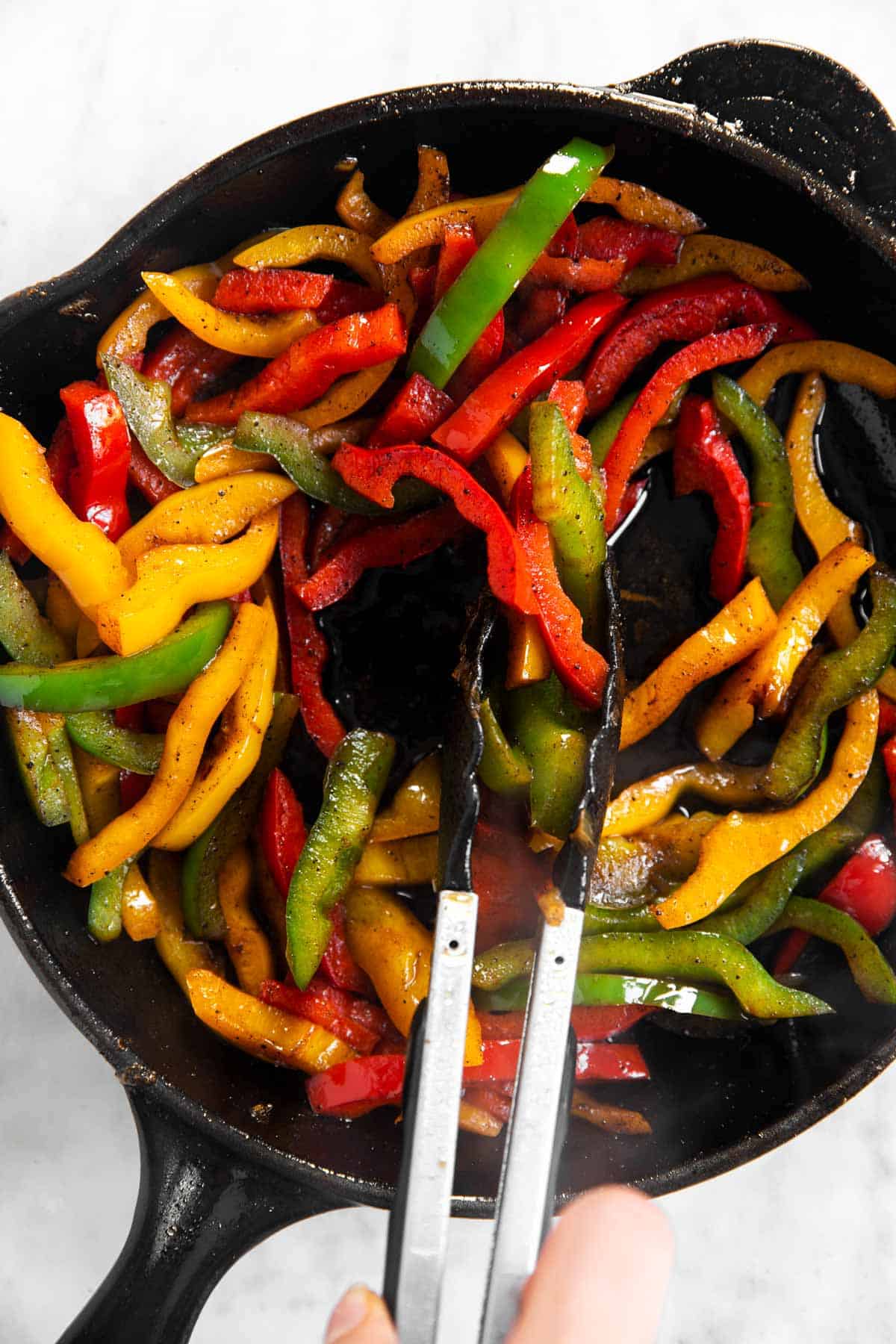 three colors of sliced bell peppers in black skillet with kitchen tongs