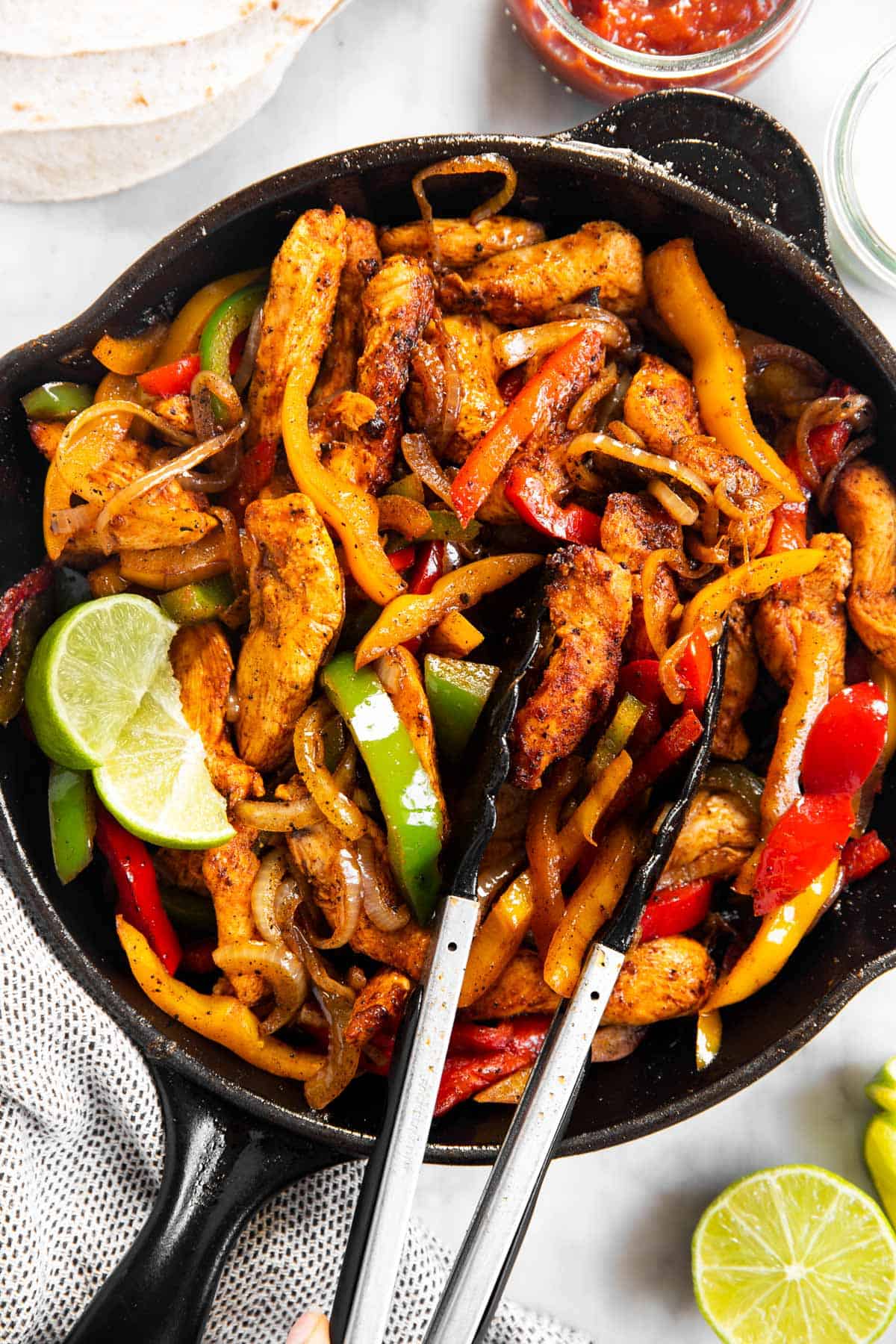 chicken fajita filling in black skillet with kitchen tongs surrounded by fajita toppings and wraps