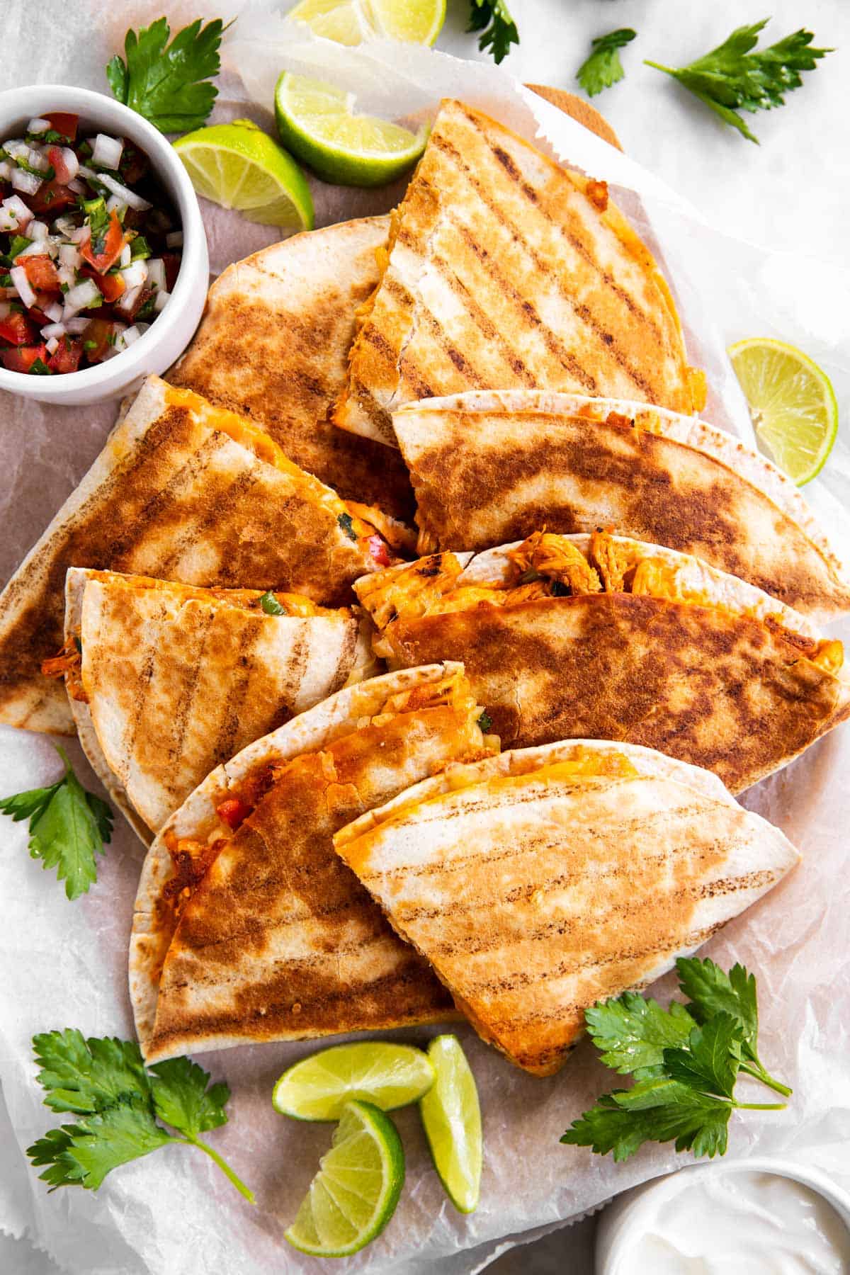 chicken quesadillas on white parchment paper surrounded by lime slices, cilantro and pico de Gallo in white bowl