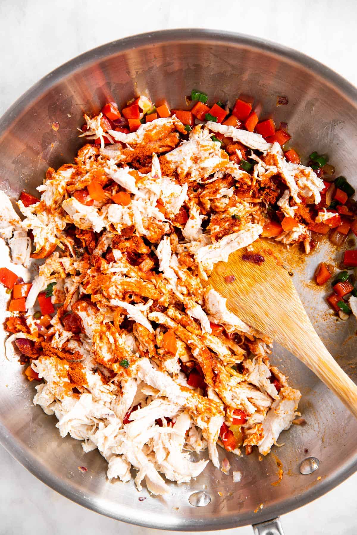 shredded chicken and seasoning being stirred into cooked onion and pepper mixture in skillet