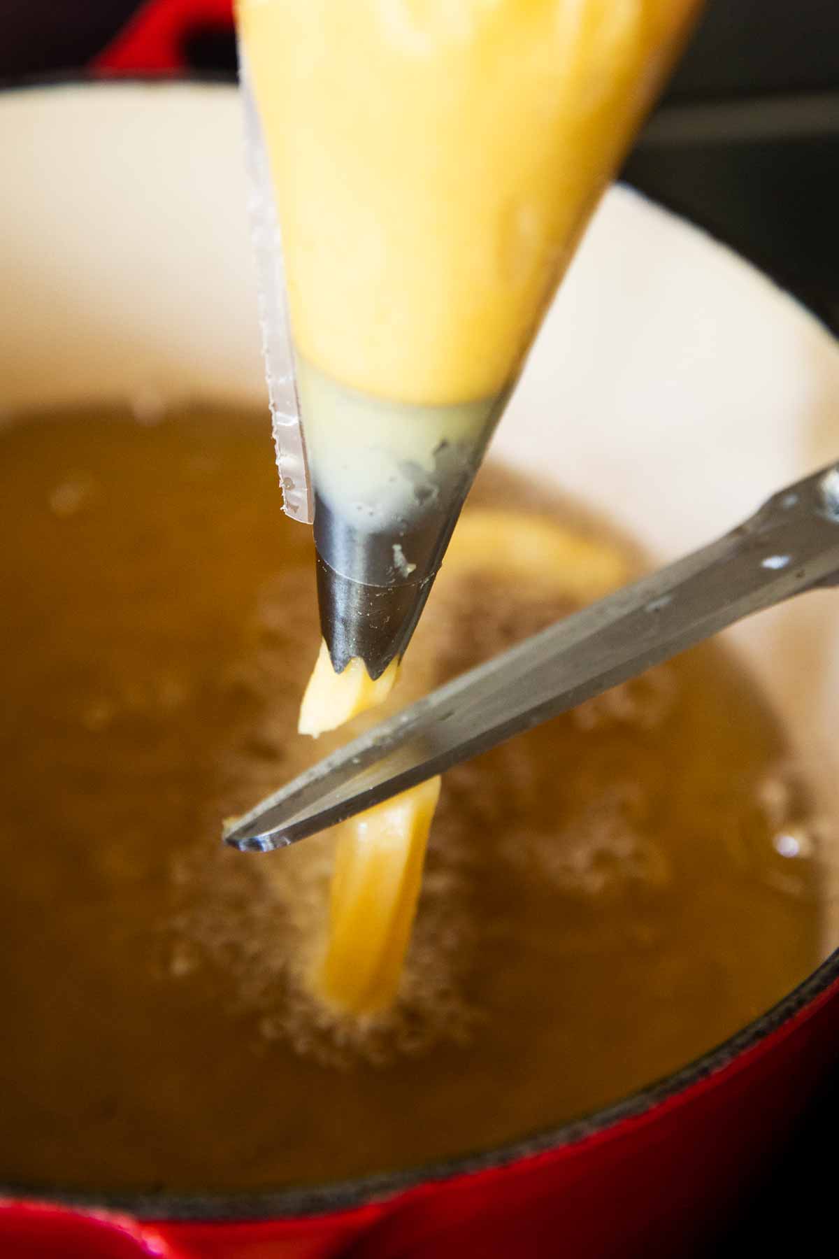scissors cutting choux pastry coming from piping bag over pot of simmering oil