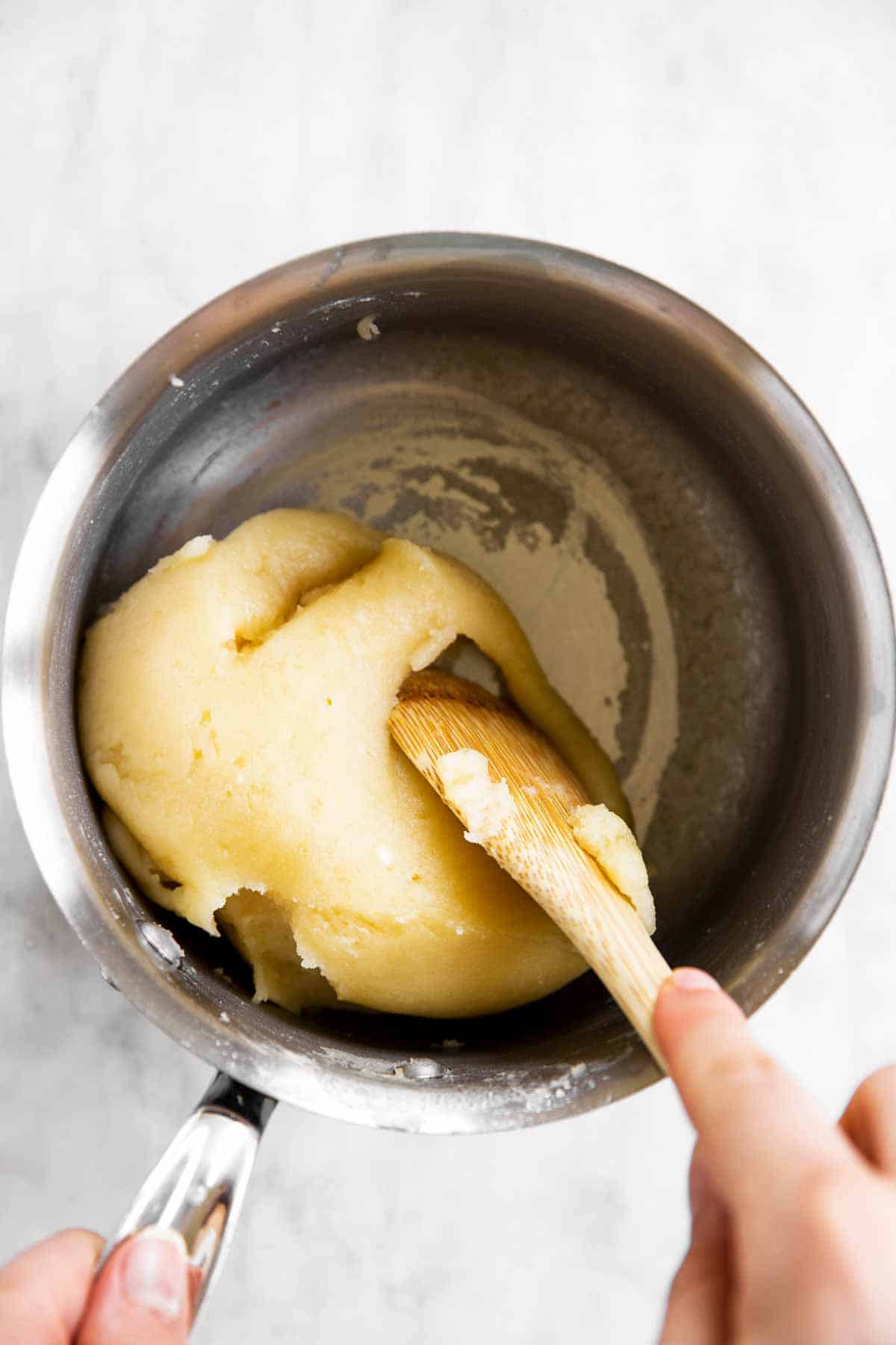female hand using wooden spoon to stir choux pastry in saucepan